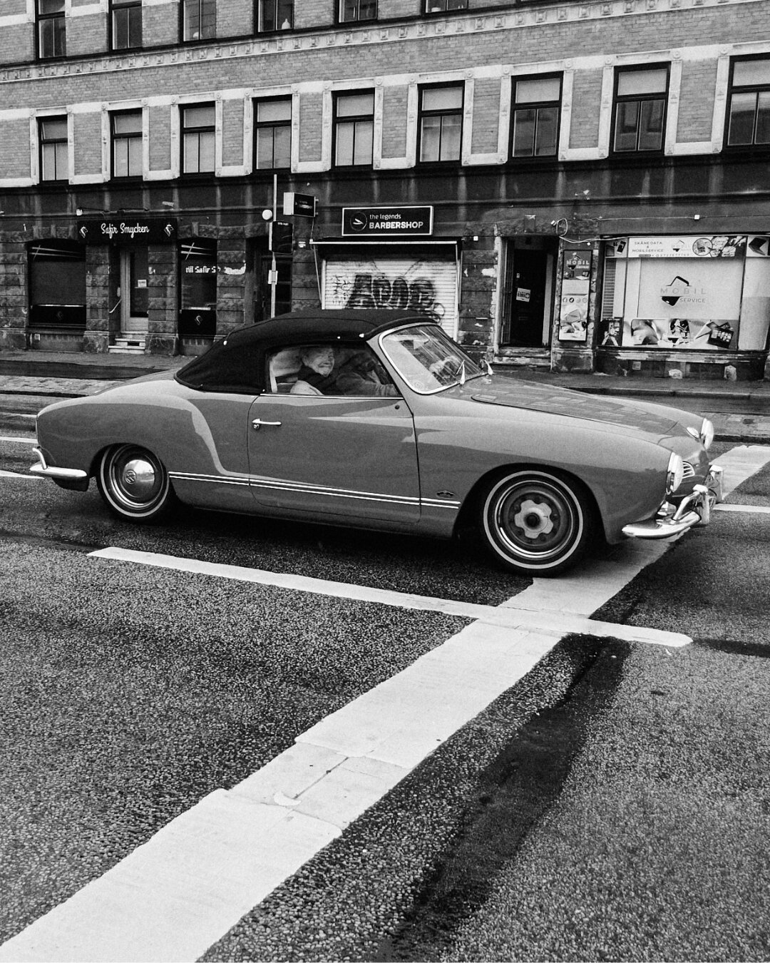 A driver inside a vintage Karmann Ghia convertible smiling through the open window on a city street.