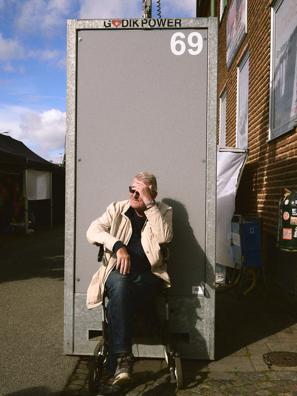 A man sitting in a wheelchair shading his eyes from the sun directly in front of a tall, gray electrical box marked "GODIK POWER 69".