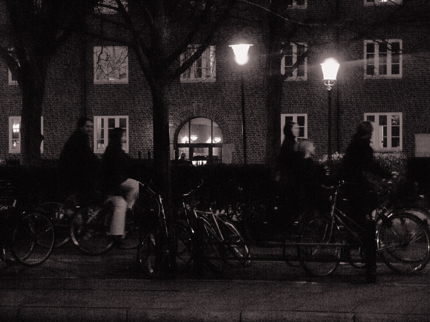 A high-contrast, grainy black-and-white night photograph of a street in Malmö. Two glowing streetlamps illuminate a brick building background while motion-blurred silhouettes of people riding bicycles pass through the dark foreground like ghosts.