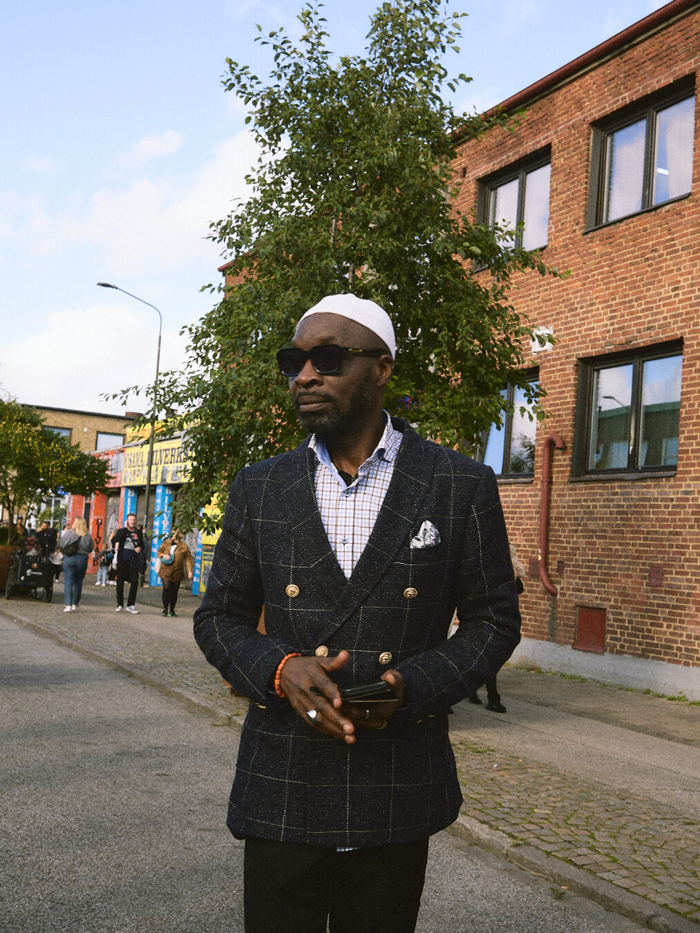 A Black man in a dark double-breasted suit jacket, white cap, and sunglasses walking forward on an industrial street near brick buildings.