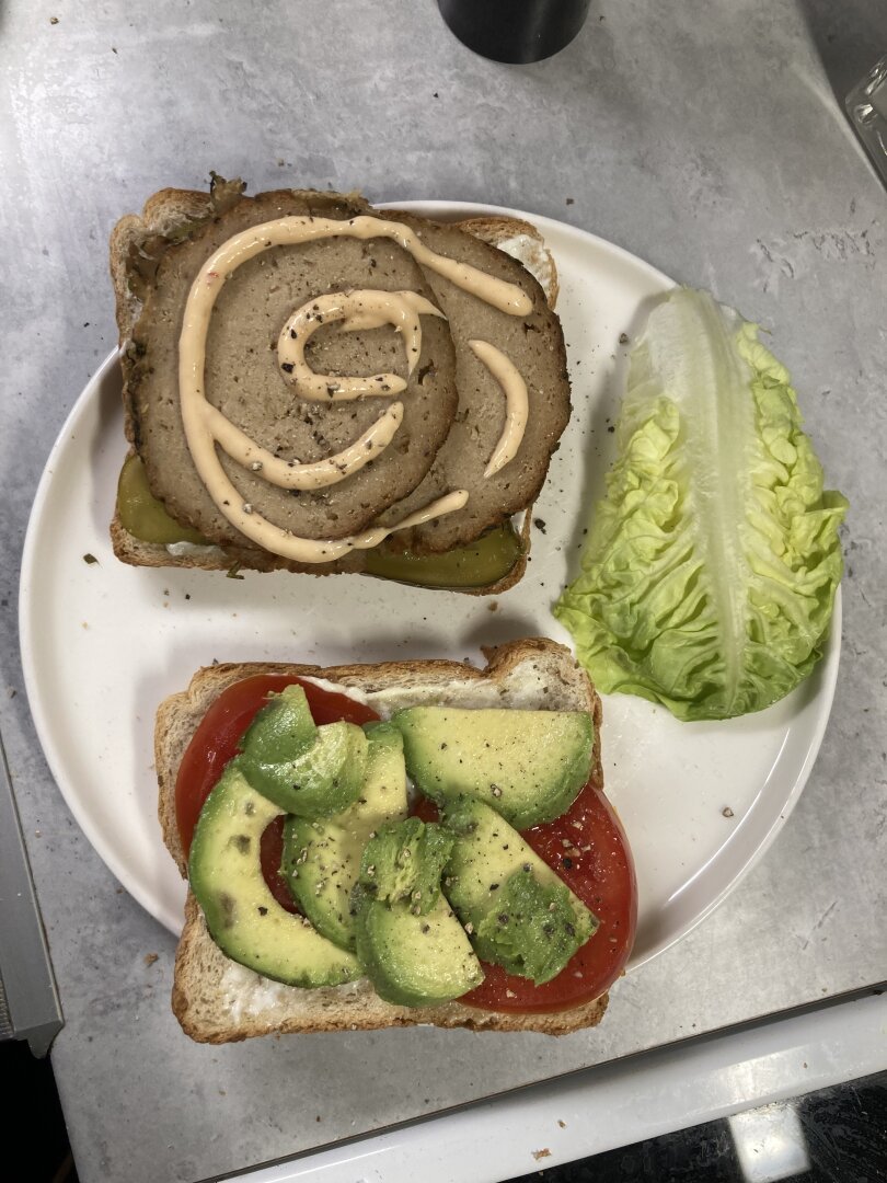 A picture of two halves of a sandwich on a plate filled with tofurky, mayo, avocado and tomato with a piece of lettuce to the side