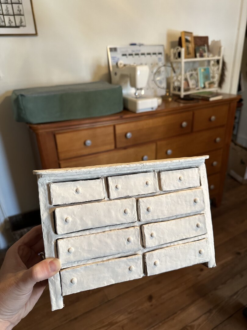 A hand holds a miniature clay dresser in front of the life size dresser