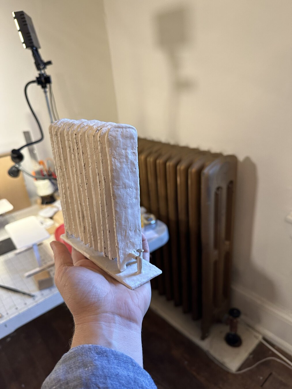 A hand holds a clay miniature radiator in front of a life size radiator