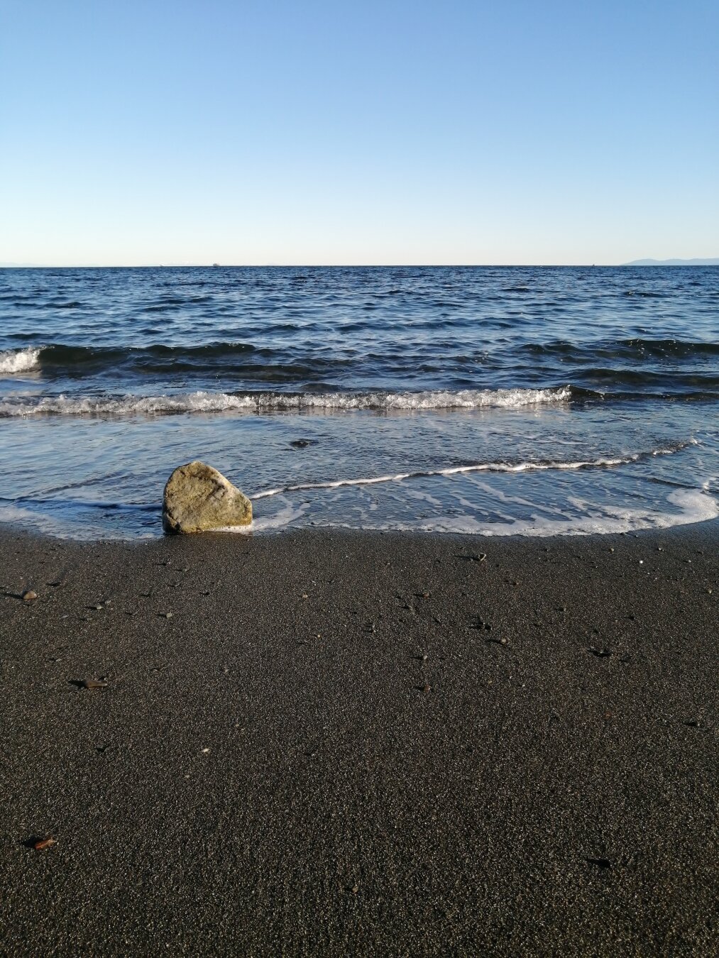 A sandy beach at low tide. The sky is blue, the sun is shining and small waves are washing over a rock at the water's edge.