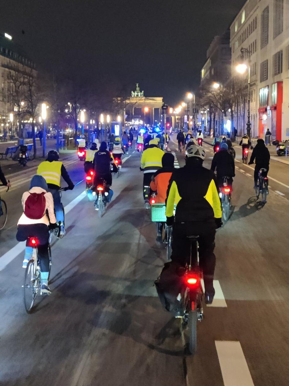 Eine große Gruppe Radfahrender bei Nacht auf Unter den Linden. Im Hintergrund ist das beleuchtete Brandenburger Tor zu sehen