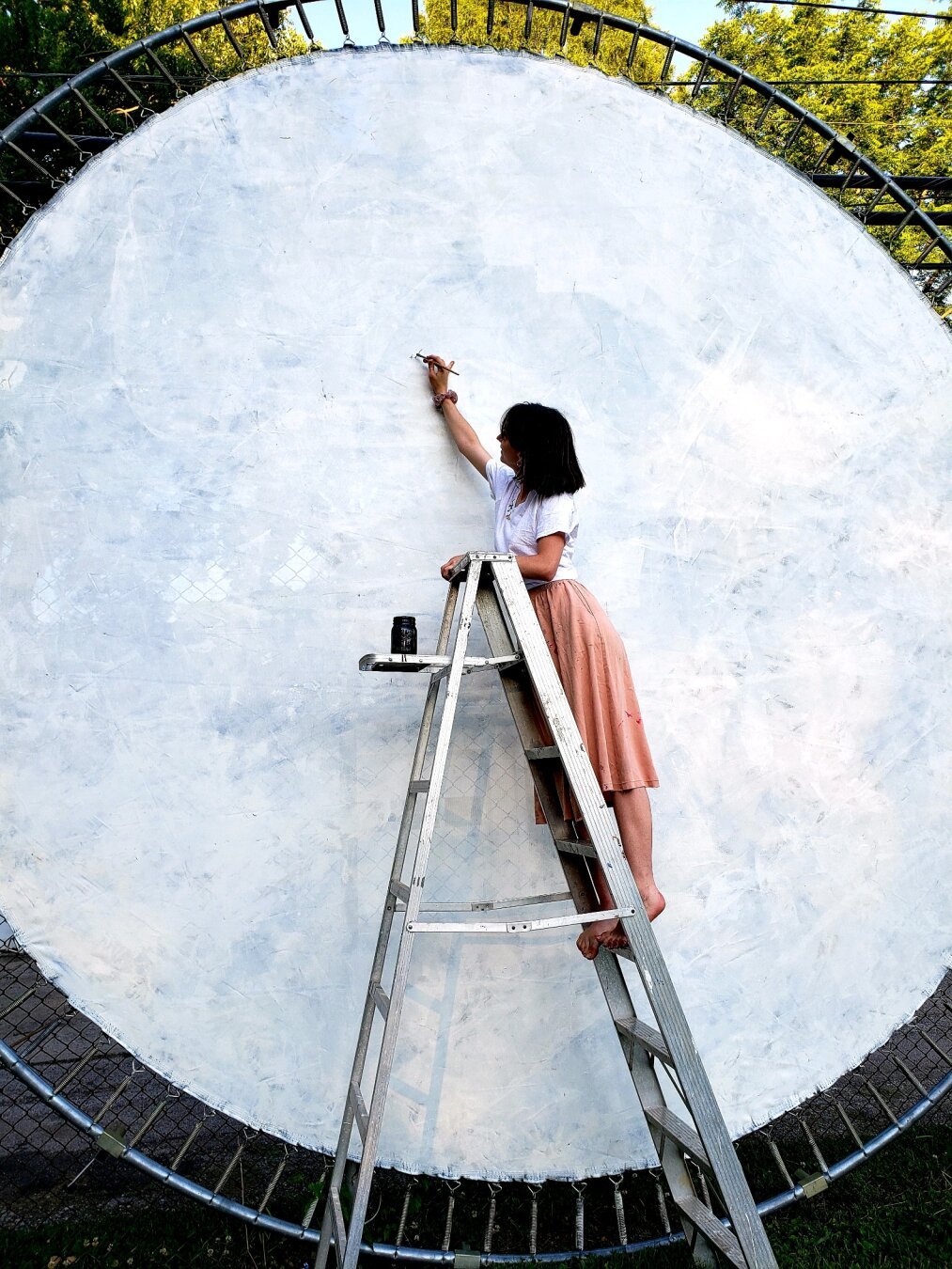 A woman standing on a ladder, painting a large trampoline that is propped on its side.