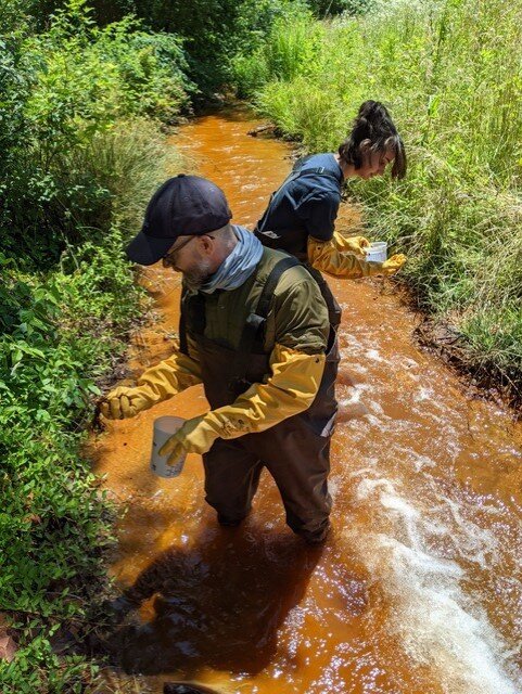 Two people wearing waders and gloves, standing inside of an orange stream, contaminated with Acid Mine Drainage.