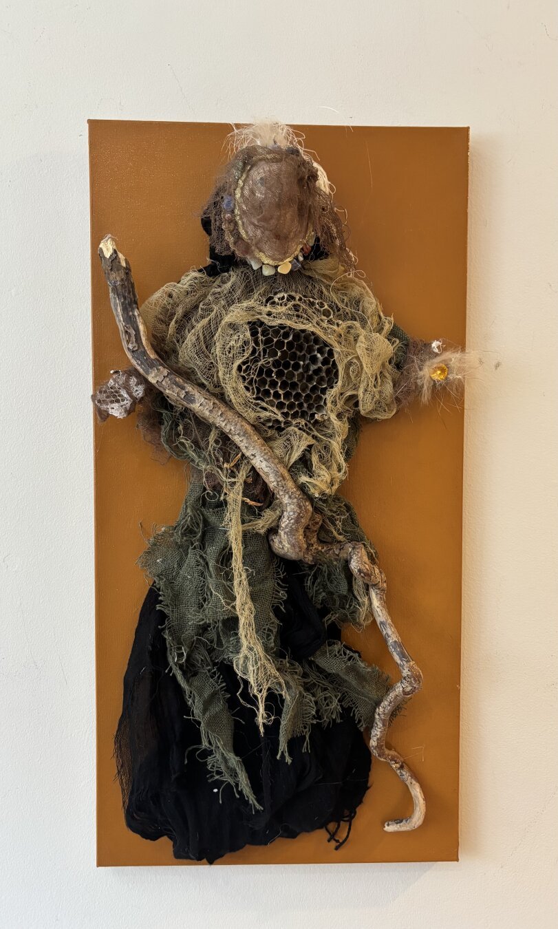 The figure of a woman dressed in earth coloured, torn and flimsy material is mounted on a dark orange canvas panel. Her face is veiled and she holds a large walking stick across her body. She stands in the traditional pose of the Virgin Mary, arms outstretched down and to the side, palms upright.