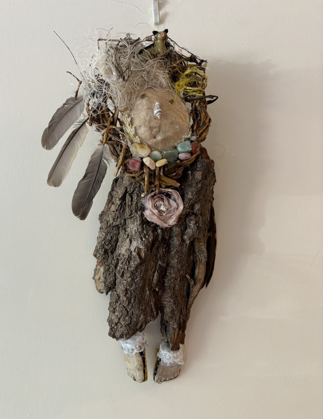 This assemblage is constructed from a piece of tree bark that resembles the shape of a human body. A paper maché mask is attached to the top. Two small stones inlaid for eyes, a tiny shell nose. The mouth is a small round opening. Hair is made from wool and loose threads. Three feathers protrude from an implied head covering on the left. The figure has clay feet and wears sandals. About 9” x 6”