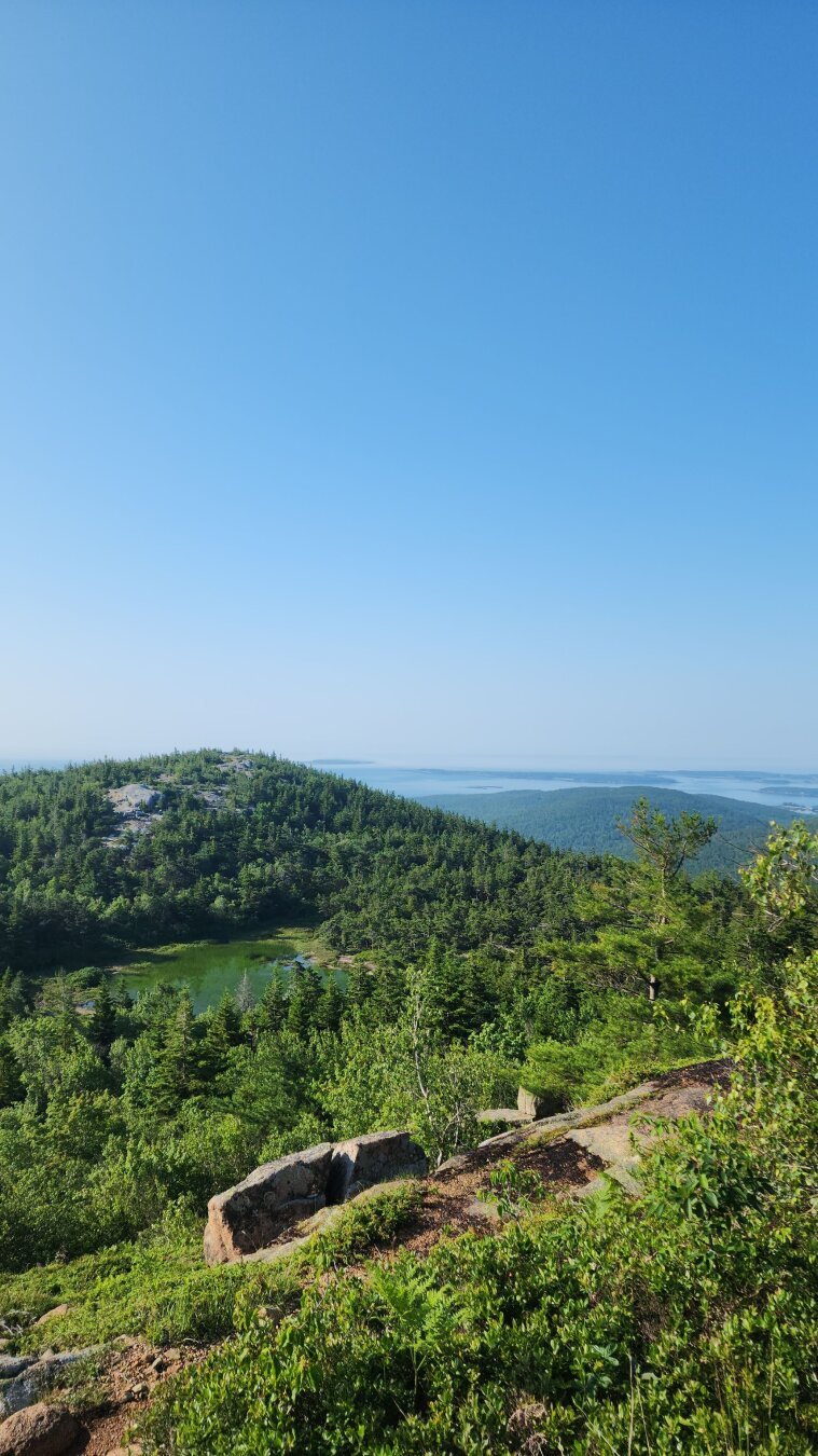 A sweeping view of the valley and green hued lake below. Taken mid way up a mountain trail. A perfectly light blue, cloudless sky behind it.