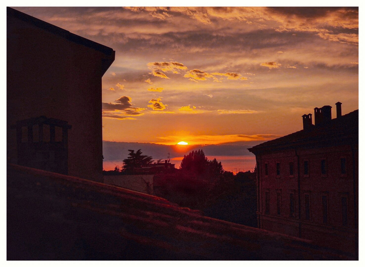 A photograph of yesterday's sunset which captures a serene sunset over rooftops. The sky has warm hues of orange, pink, and purple, with scattered clouds. There are silhouetted buildings and trees frame the scene.The foreground includes a stone wall and the dark outline of a building. A photograph of yesterday's sunset which captures a serene sunset over rooftops. The sky has warm hues of orange, pink, and purple, with scattered clouds. There are silhouetted buildings and trees frame the scene.The foreground includes a stone wall and the dark outline of a building.