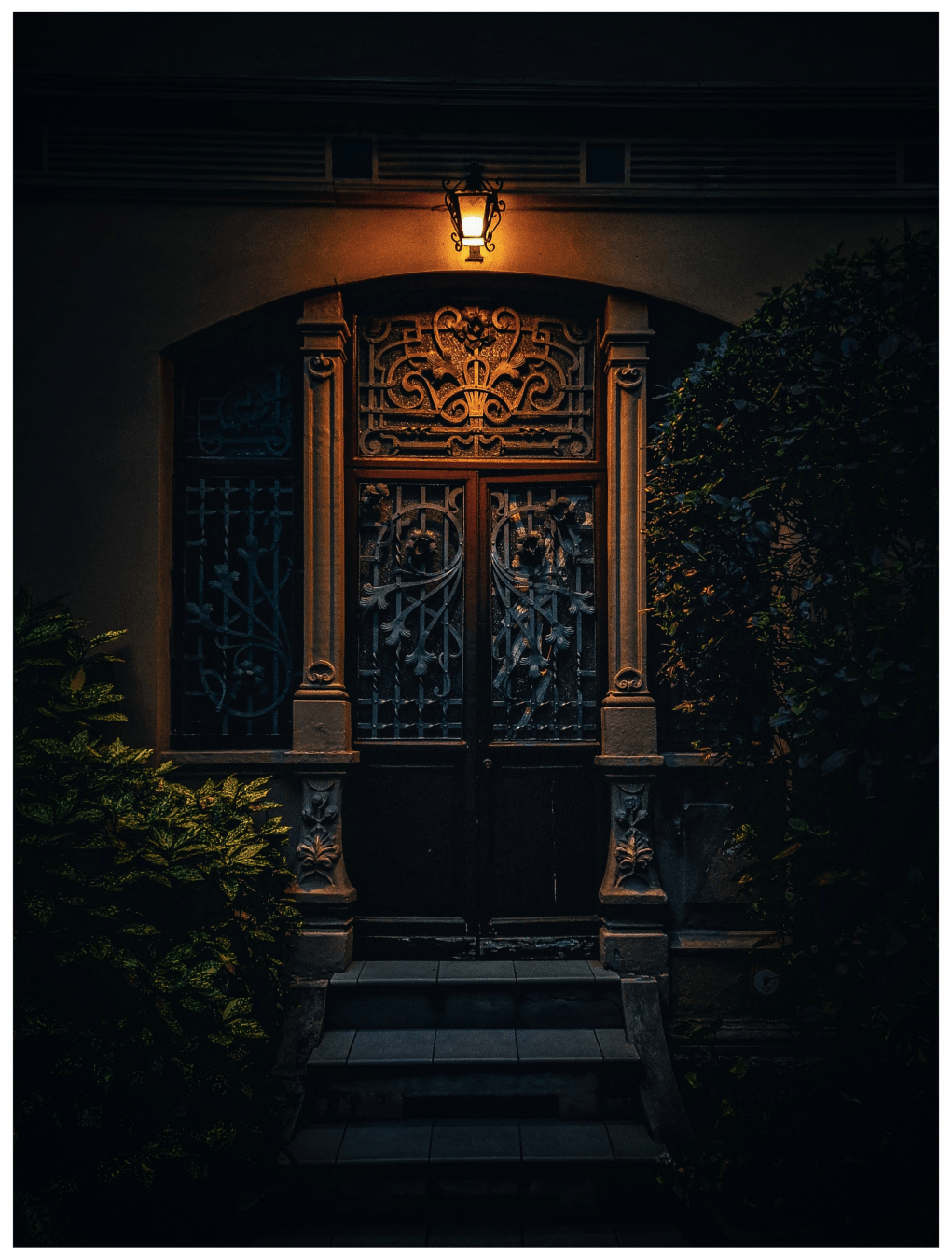 A photo of a doorway at late evening. The doorway is ornately framed with stone and metal detailing, and features a partially visible wooden door with intricate wrought iron metalwork. There isa small, lit lantern which hangs on the wall above the door. The steps leading to the door are visible, and lush green plants frame the doorway on either side. A photo of a doorway at late evening. The doorway is ornately framed with stone and metal detailing, and features a partially visible wooden door with intricate wrought iron metalwork. There isa small, lit lantern which hangs on the wall above the door. The steps leading to the door are visible, and lush green plants frame the doorway on either side.