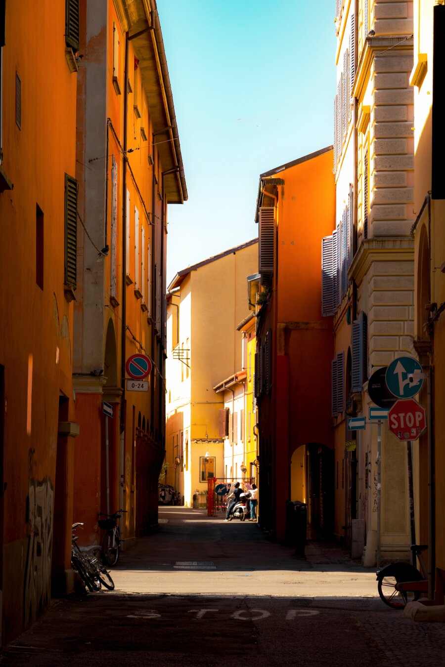 This is a photo of Via Paglietta. It Is a narrow, sun-soaked street framed by tall, vibrant orange and yellow buildings. Some sunlight casts warm highlighys and deep shadows along the street. There are some street signs like a red stop sign and a no-parking sign mounted on the building walls. Near us, there are parked bicycles which lean against the bases of the houses. Further down the alley though there is a person on a scooter with another about to climb on. The sky above is nice and clear blue.