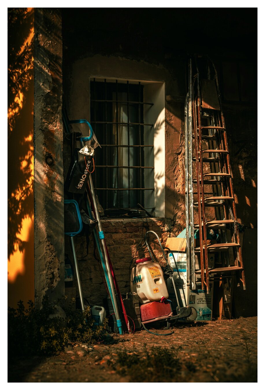 This photo is of a friend's outdoor storage area (it's under an arch) against the exterior wall of a building. On the left-hand side, there are various long-handled pool cleaning nets and tools which lean against yellow wall dappled with leaf shadows. Behind them to the right-ish is a barred window. Underneath the window there is a a few things a red and white portable sprayer tank and boxes etc. on the right there are two tall ladders. One is metal and one is wooden, they are leaning on the weathered stone building. The photograph is filled with warm evening sunglight and a collection of items of a life filled with love.