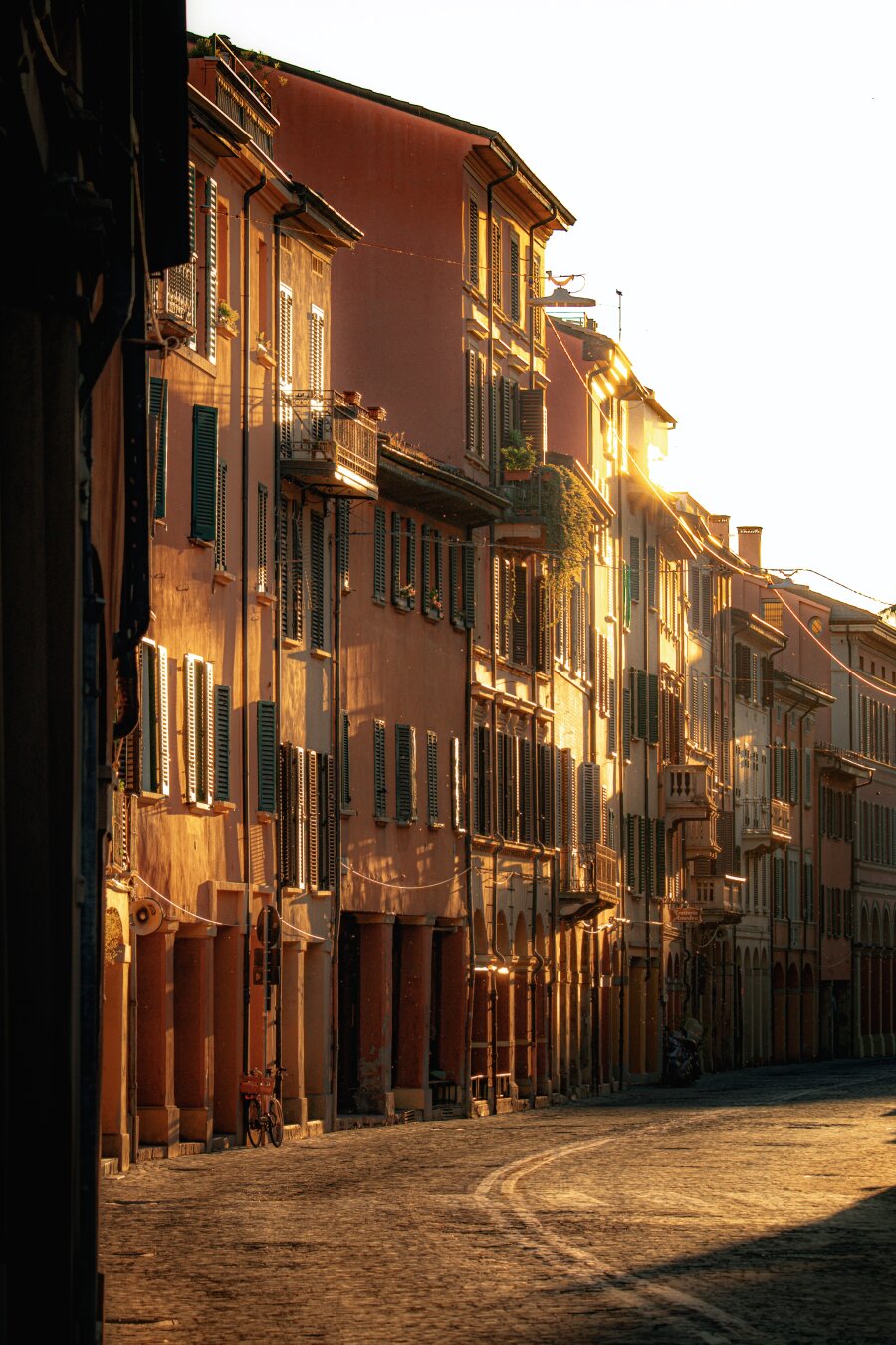 This is a photography of a street in Bologna (via Saragozza). It Is a cobblestone street which curves between tall buildings painted  shades of terracotta and yellow. There is some golden sunlight which streams down the street, illuminating the building faces, shutters of the buildings. On the left there is a bicycle against the wall near an arcade.