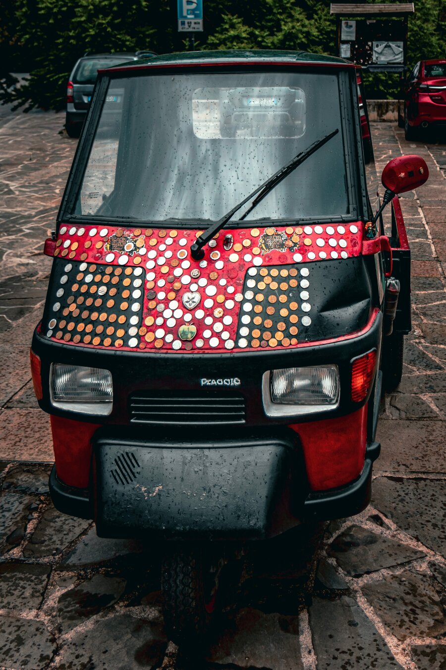 A photography of a really cool red Piaggio Ape 50 which has lots of coins symbols and knick-knacks stuck to it. It's in a car-park.