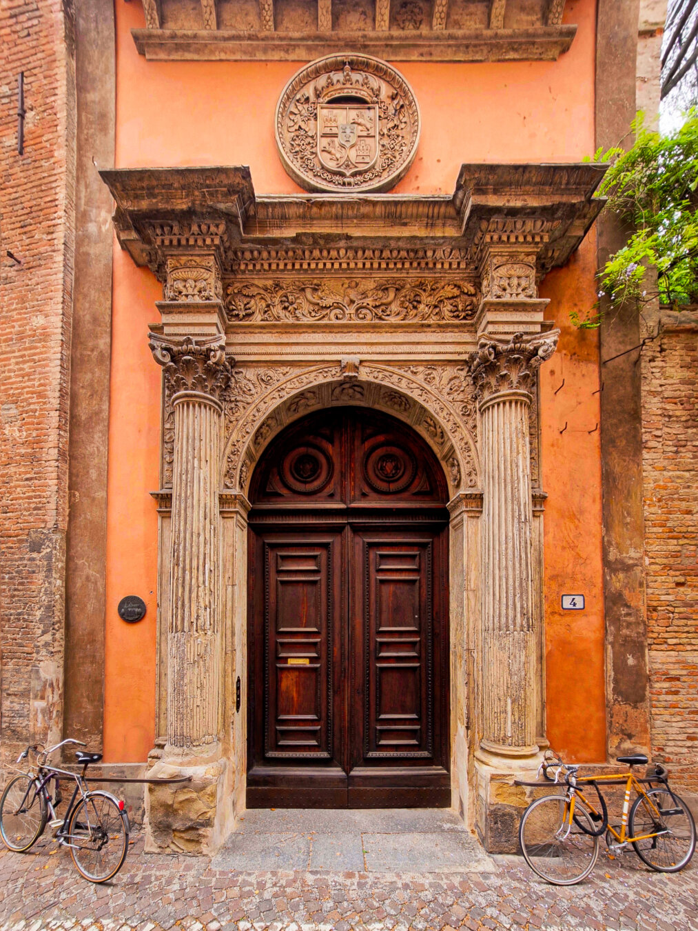 This is a photo of the main entrance to Collegio di Spagna. It is pretty big, with ornate stone columns and relief which framedouble dark-wooden doors that are set into a terracotta-orange building facade. The columns are Corinthian and hold up the intricately carved stone frieze/relief and a circular stone crest mounted just above. Two bicycles are parked on the cobblestone street in front of the entrance, one on the left and a one on the right.