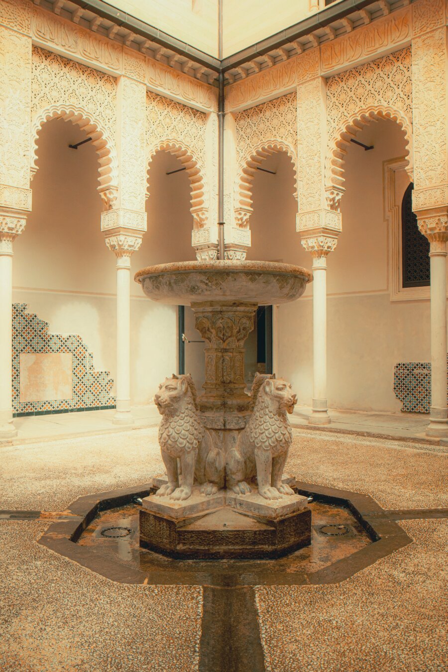 The photograph is of the stone fountain sitting at the centre of an open-air courtyard styled with Moorish architecture which is located in Rocchetta Mattei. The base of the fountain has lion statues supporting a big central basin inside an octagonal pool. Behind it in the courtyard are intricately carved arches supported by columns. There is also decorative tilework lining parts of the lower walls.