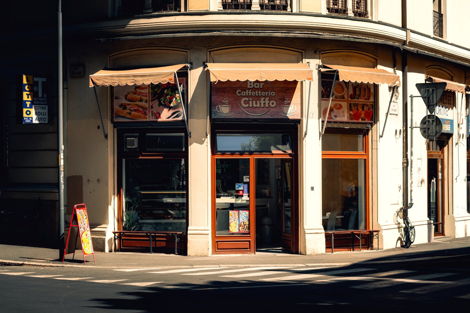 This photo is of the outside of "Bar Caffetteria Ciuffo", on a sunny street corner. It has fabric awnings above the large glass windows and an open central glass door. On top of the doors and windows there are signs with the café's name and on either side has pictures of some of the products. On the left-hand side there a vertical yellow "LOTTO" sign hanging on the wall and a red A-frame (? Is that what it's called?) menu on the footpath.