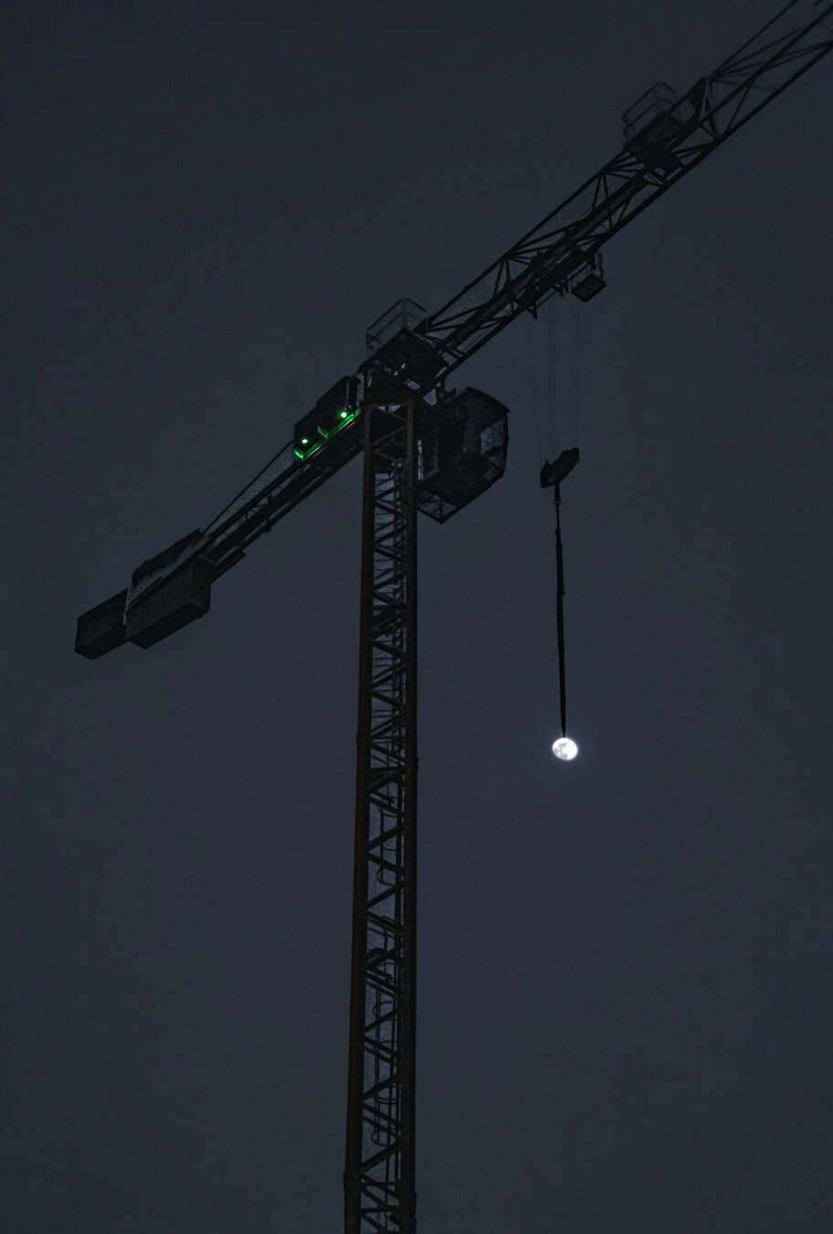 The photograph is of a construction crane in Piazza San Francesco silhouetted against a dark sky. The crane's metal structure has a lot of dark lines and angles. There are two green lights glow on the crane's arm. On the right-hand side,  hanging from a cable on the crane's arm is the moon. (It's not really hanging, I just positioned it that way with perspective!)