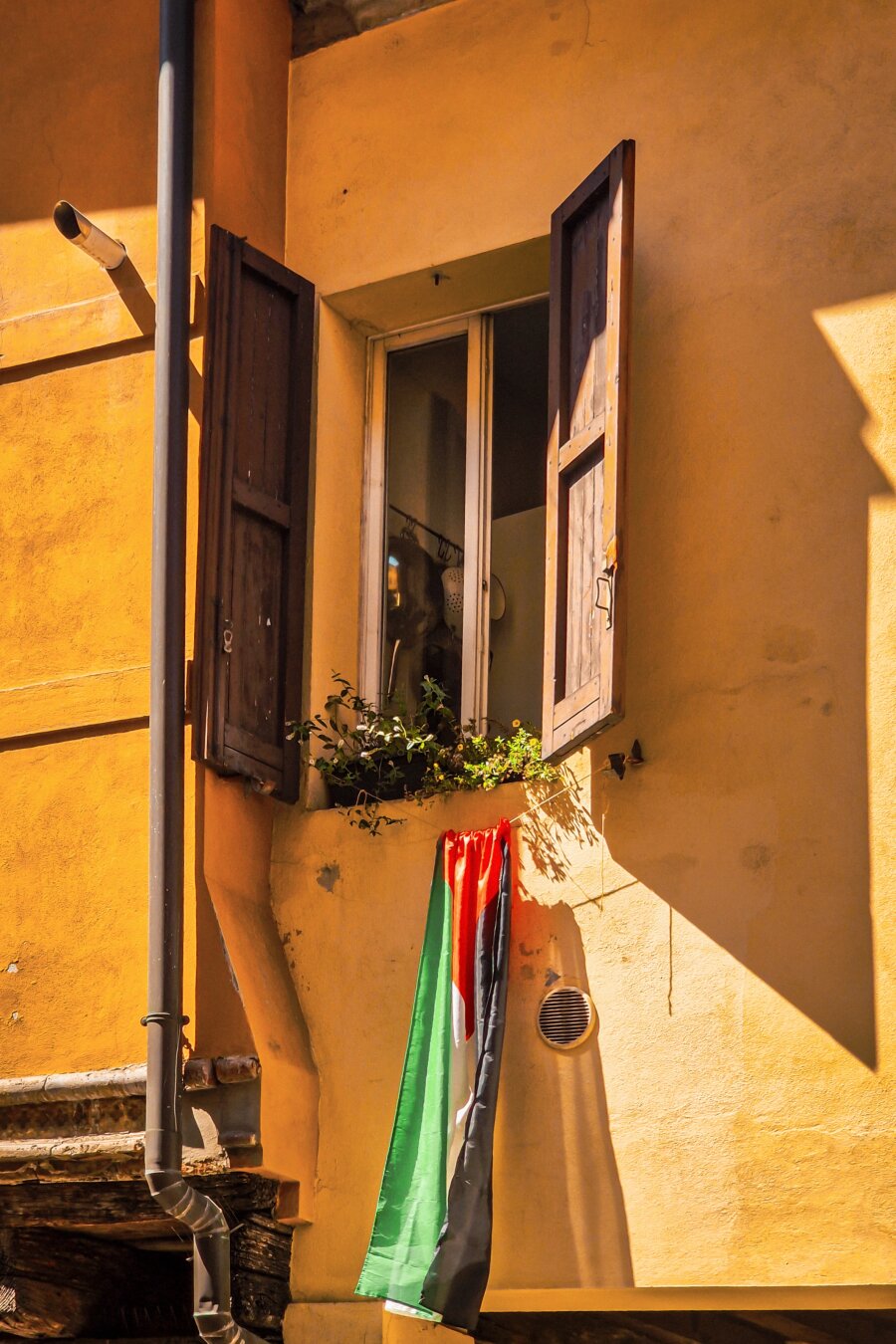 This photo is of a window with a  Palestinian flag hanging underneath it. The window has dark wooden shutters on a yellow building exterior. There is some potted plants on the windowsill, while a dark drainpipe runs along the wall to the left. Some bright sunlight casts strong shadows across the warm yellow, textured plaster wall.
