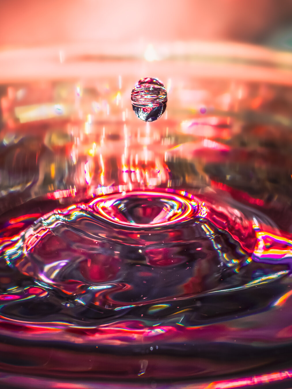 This is a macro photograph of a  water droplet suspended momentarily in mid-air above a ripple pattern underneath in a wine glass. There are vibrant red, pink, orange, and golden colours which reflect through the water surface, creating a trippy, kaleidoscopic effect. The droplet  is sharply focused (thankfully 😂) and is mirroring the surrounding colourful lights against a soft bright background.