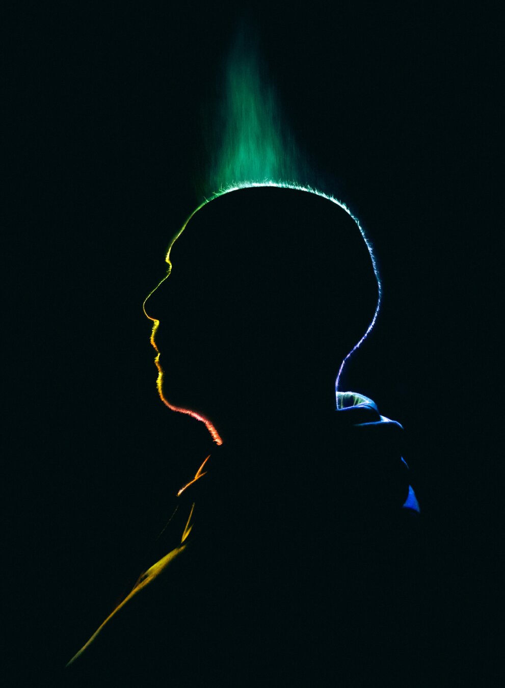 This photo is of my partner Peter. It is mostly black but because of the lighting you can see the outlines of his side profile and his hoodie. The lighting is in rainbow formation and goes from yellow-orange at the front (or left hand side) to blue/violet at the back (or right hand side).
At the top of his head there is a triangular shaped green patch which is a combination of the fallen water droplets and the led light.