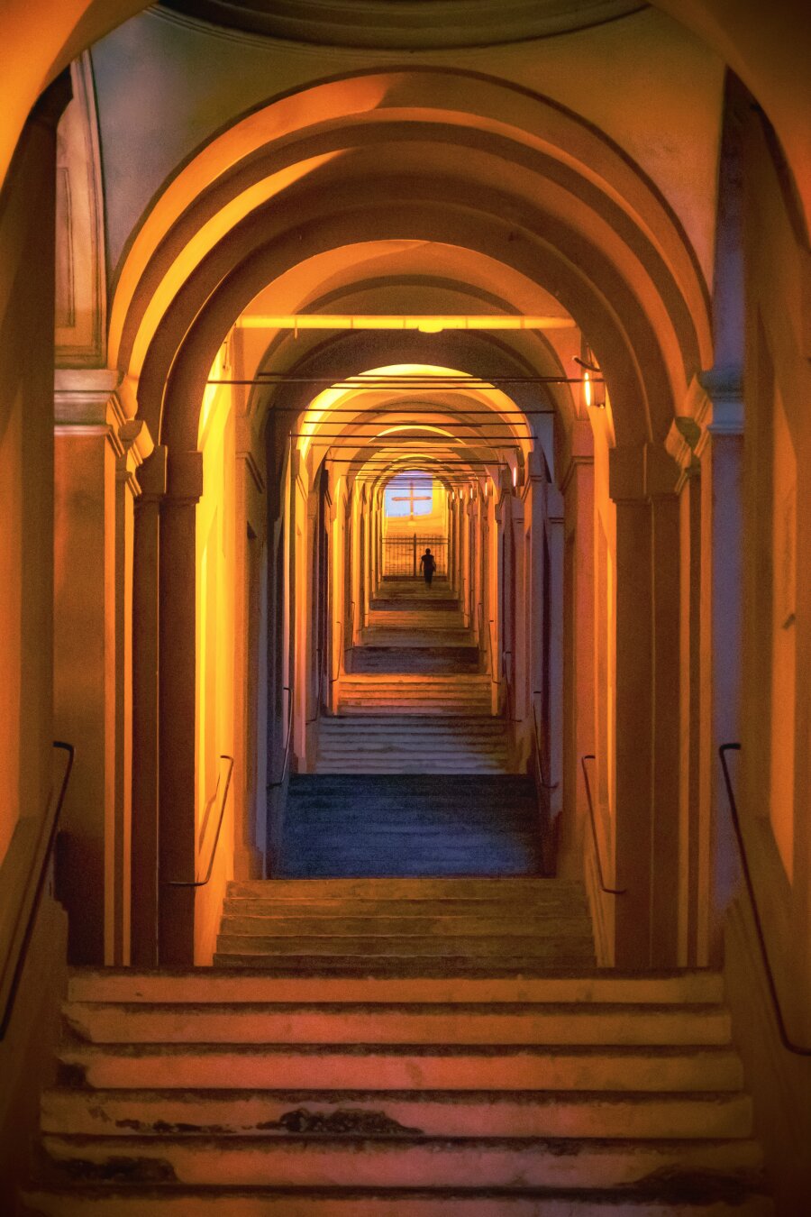 This photo is of a long symmetrical portico which is framed by  repeating rounded archways. They are lit with a warm, golden glow from the lights. There are flights of stone steps that lead upwards through the porticoes toward a distant opening (which is actually gated because it's about half six in the morning 😂). In the centre, up the steps there is  a lone silhouette walking towards the gate.