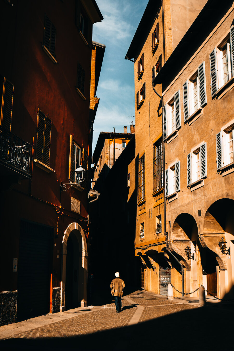 A photograph of a sunlit, narrow Bolognese alleyway with tall, old brick buildings on either side. (Via de' Pepoli) A solitary person with a white hair and a light brown coat walks away from the camera - they look very small compared to everything. The buildings have shuttered windows and wrought-iron balconies. Shadows cast long, dramatic lines across the cobblestone street. The sky above is clear and blue with some wispy clouds.