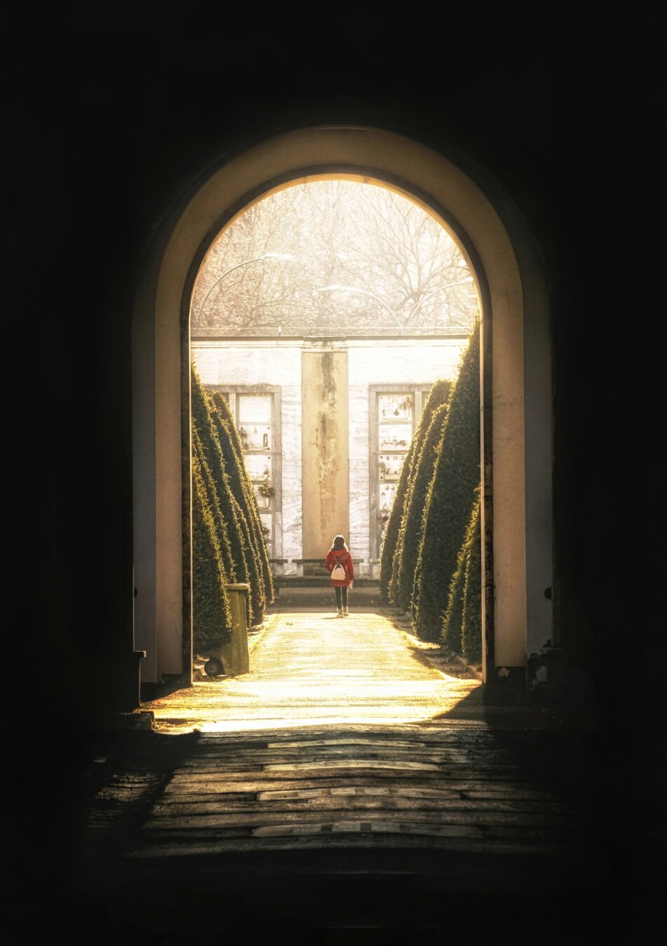 The photograph from La Certosa cemetary. It is of a view looking out from a dark, shadowed archway into a brightly lit outdoor area. There is a single person in a red coat and backpack walking away in the distance along a central pathway illuminated by soft, golden light. The path is bordered on both sides by conical green hedges leading towards a light-coloured building.