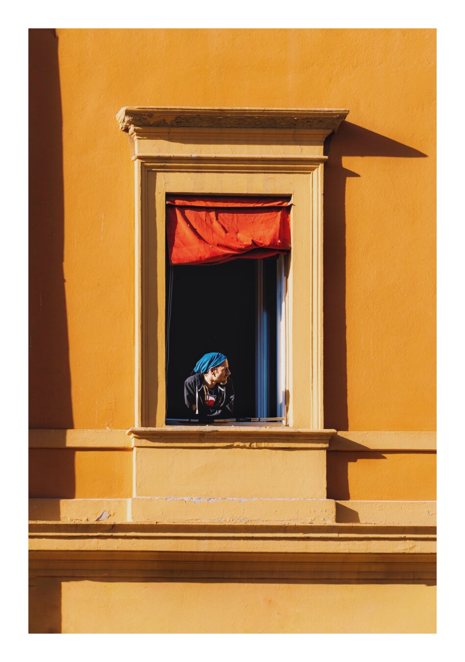 This is a photo of a  person wearing a bright blue head scarf as they lean out from an open window of a bright yellow building. They are looking towards the right hand side.  Above them there is a red window blind bunched up and inside the upper frame of the window. The sunlight highlights the wall and creates shadows from the cornices/ledges of the window.