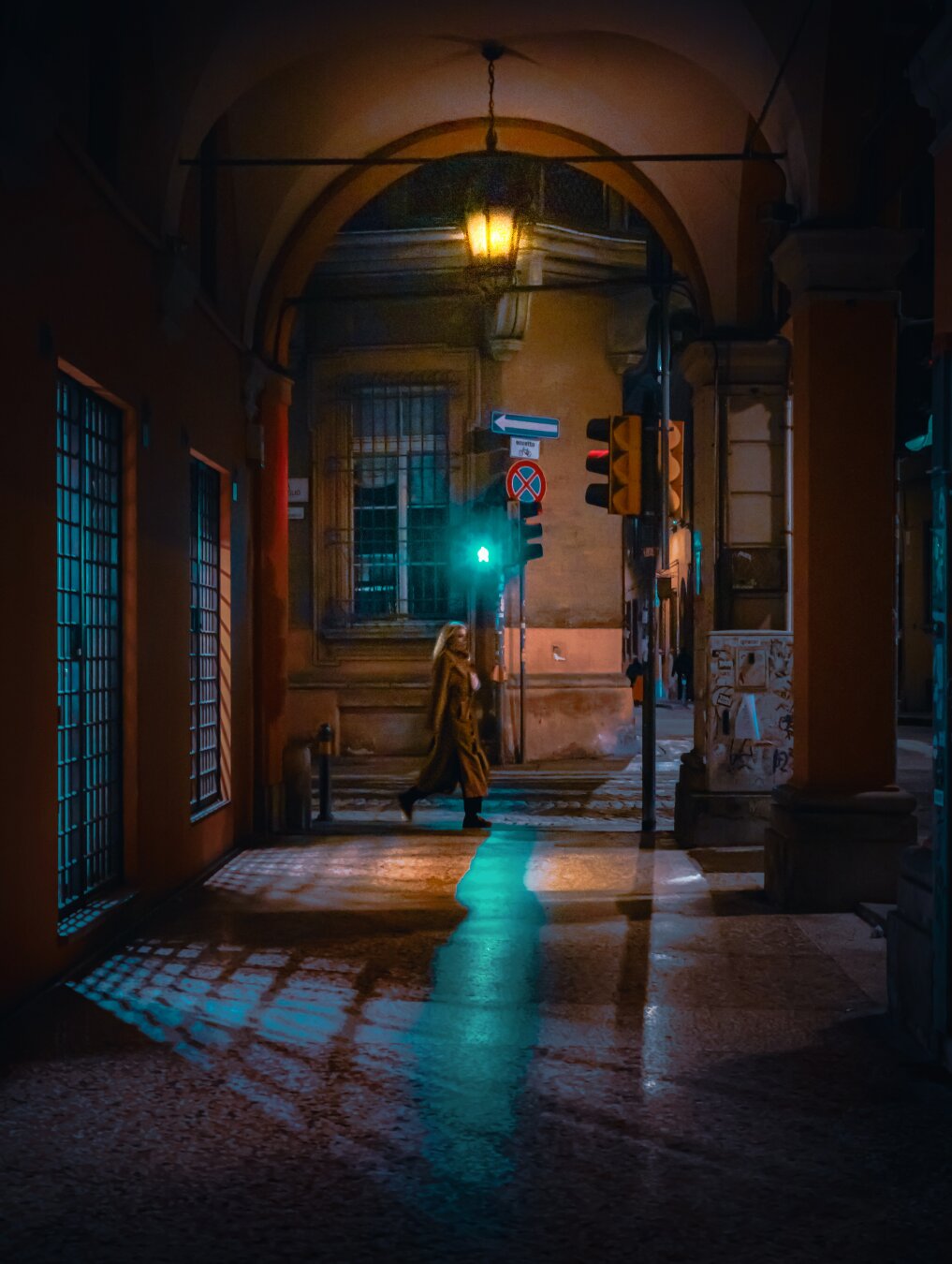 A photo of a nighttime urban scene, focusing on a crossing bathed in green light coming from a traffic light. A woman is captured mid-stride as she crosses the street, her brown long coat flowing behind her. The traffic light is prominently displayed, showing a bright green light. Overhead, the crossing is framed by a series of arches, typical of Bologna A vintage-style lantern hangs from the archway, casting a warm, yellow glow that contrasts with the cool green light below. The buildings on either side of the crosswalk are aged, with barred windows..The ground is paved with stone blocks, reflecting the green light.