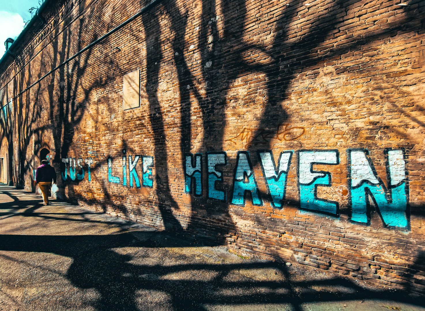 A photograph of a sunlit brick wall with bold, blue and white graffiti which says "LIKE HEAVEN." The wall casts shadows of tree branches, creating a textured pattern. A person with brown hair, wearing a purplish red jacket and beige trousers, walks along the footpath on the left.