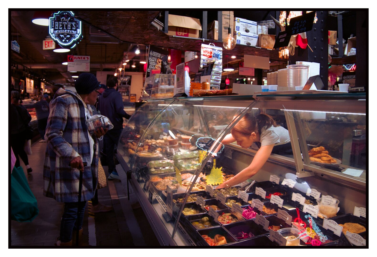 Color photograph taken in Reading Terminal (in Philadelphia).

A woman reaches into a large glass display case to pull something out for a customer, the lights inside the case illuminating her as her customer looks on from the other side.