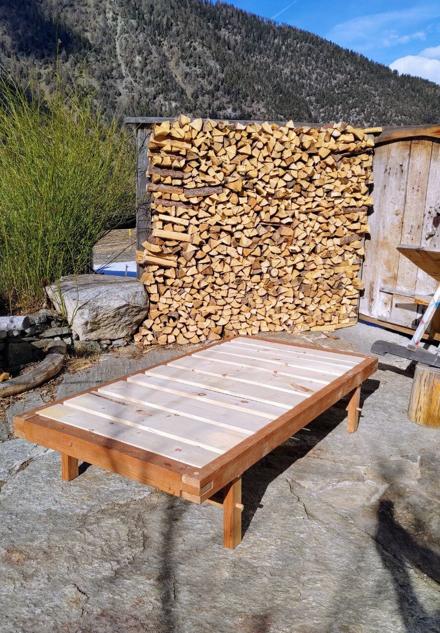 wooden bed on stone square with firewood and door in the background