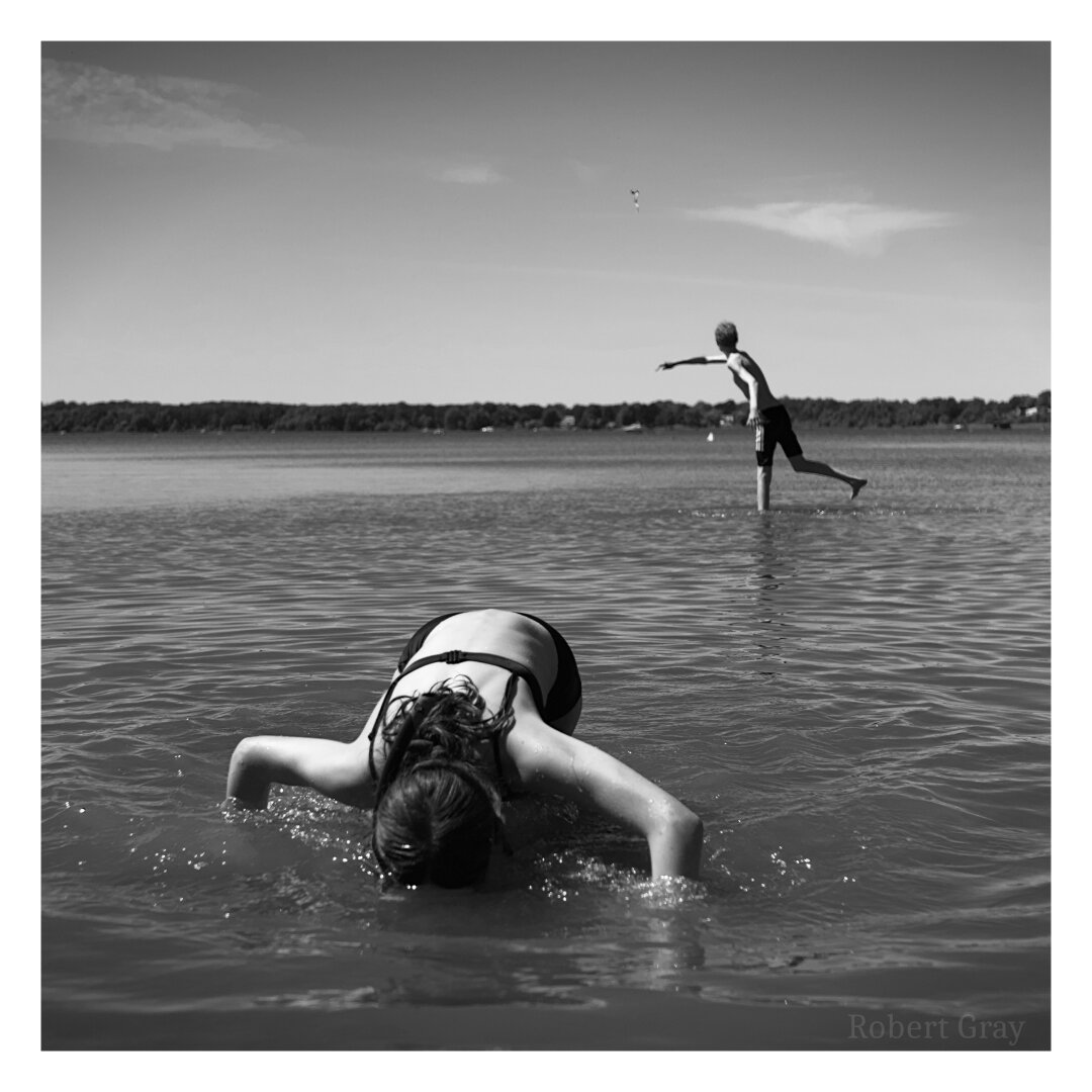 Monochrome image of a girl dipping her face in the water. In the background and further from the shore is a boy wearing trunks who is throwing an object away from the camera.