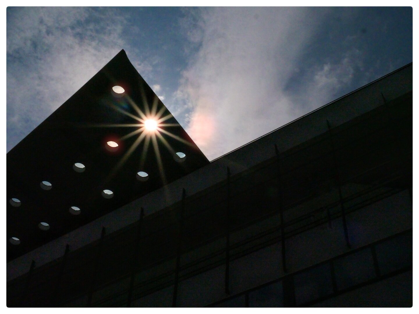 View from the ground looking up at an angle to a roof structure which juts out over the street and which has about ten holes in it. Through one of the holes a sunburst comes through creating some lens flare. The scene is overlaid with the outline of another building, creating a form like two mountain peaks next to each other.