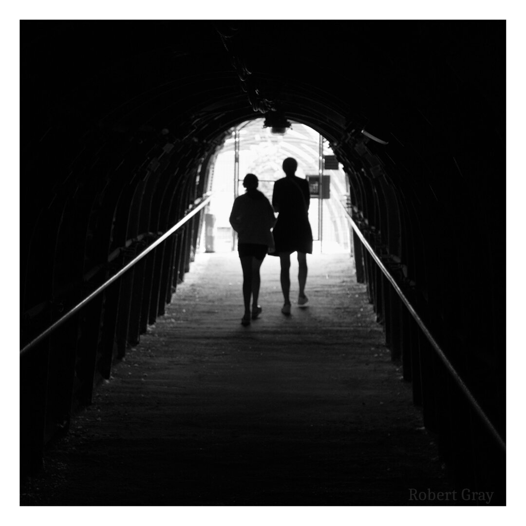 Two distant figures, a woman and a girl, are seen in silhouette from behind as they walk out of a dark tunnel into bright sunlight.