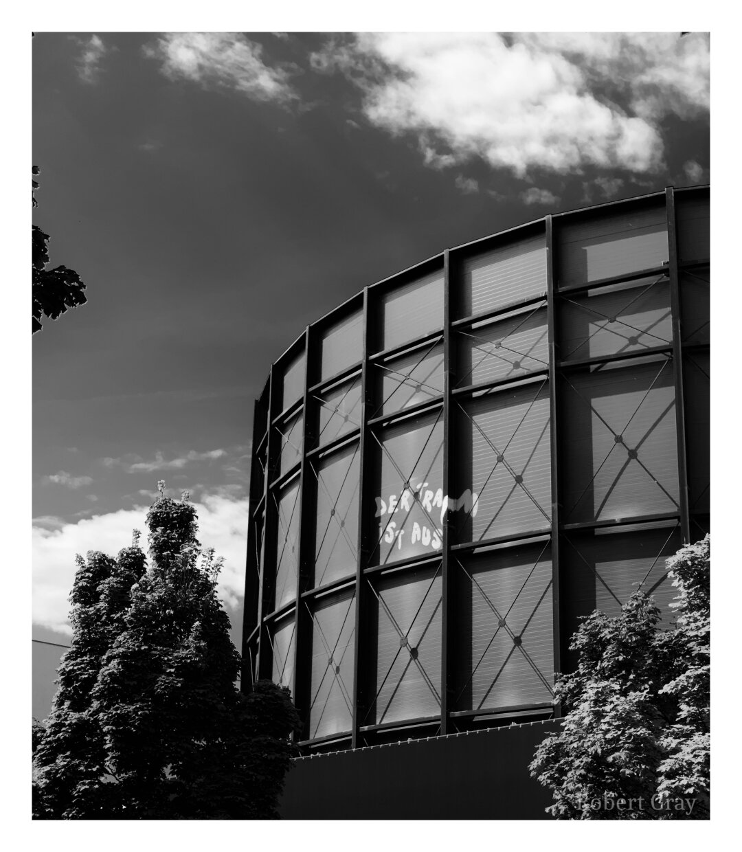 A monochrome image of a gasometer with the text "Der Traum ist aus" ("The dream is over") painted onto one of its panels. The image is high-contrast and the tone sky has been adjusted to make it appear very dark with white, fluffy clouds.