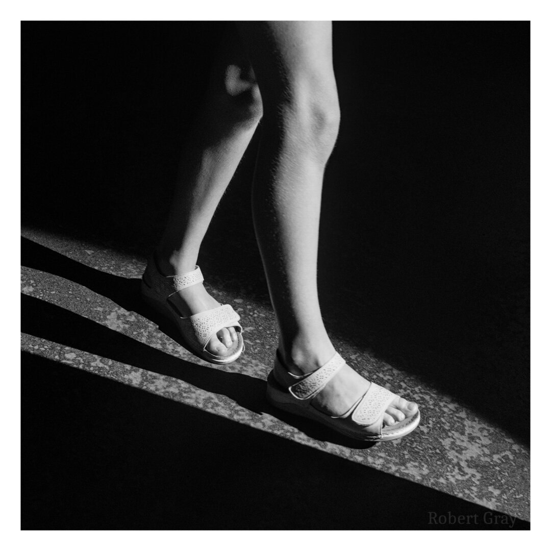 A girl with bare legs and wearing white sandals walks through a patch of light in an otherwise dark room. Only her lower legs and sandals are visible. The image is in high contrast monochrome.
