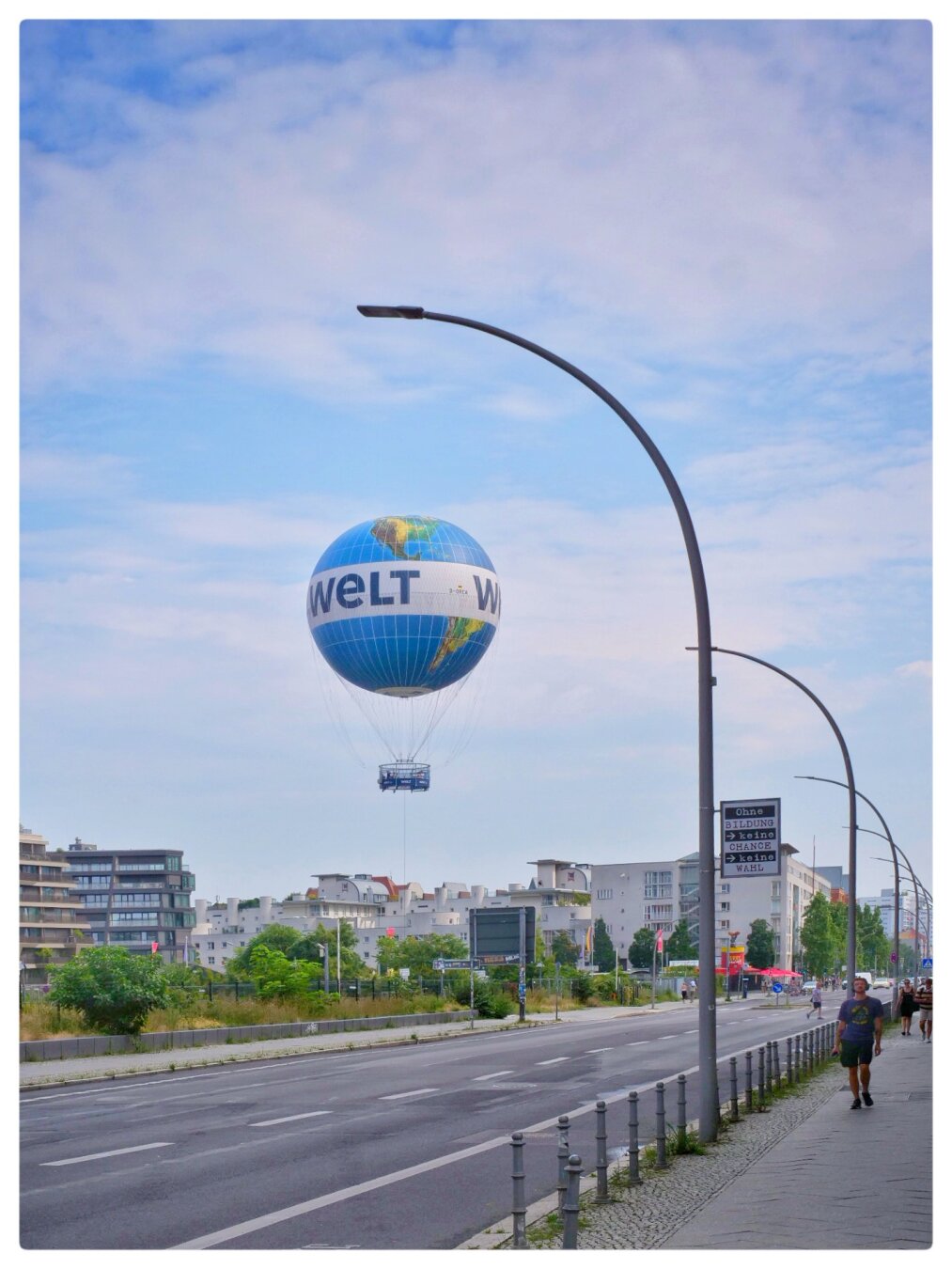 A hot air balloon carrying the text „Welt“ rises up into the sky over balloon. It is tethered to the ground by a cable. The shot is framed by a street lantern with a curved shape, echoing the rounded form of the balloon. An empty street runs diagonally through the scene from the bottom left.