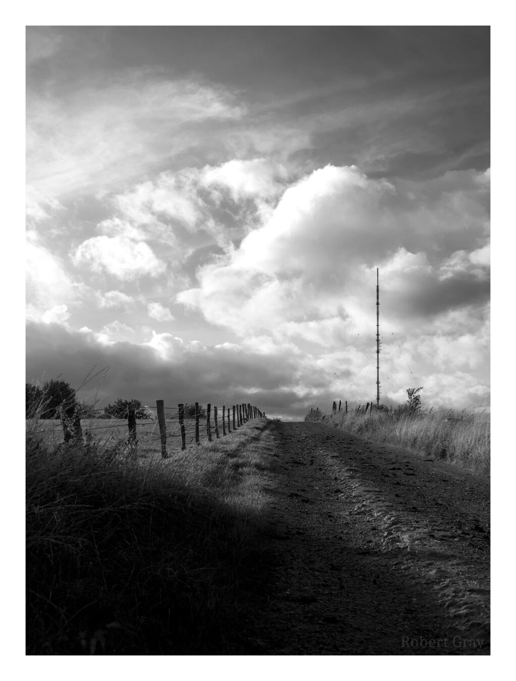 A gravel track leading away from the viewer and slightly uphill. To the left there is a simple farm fence. On the brow of the hill is a mobile phone mast secured with steel cables running at a diagonal to the mast. The sky is dark and dense with full, angry-looking clouds, as if a storm is brewing.