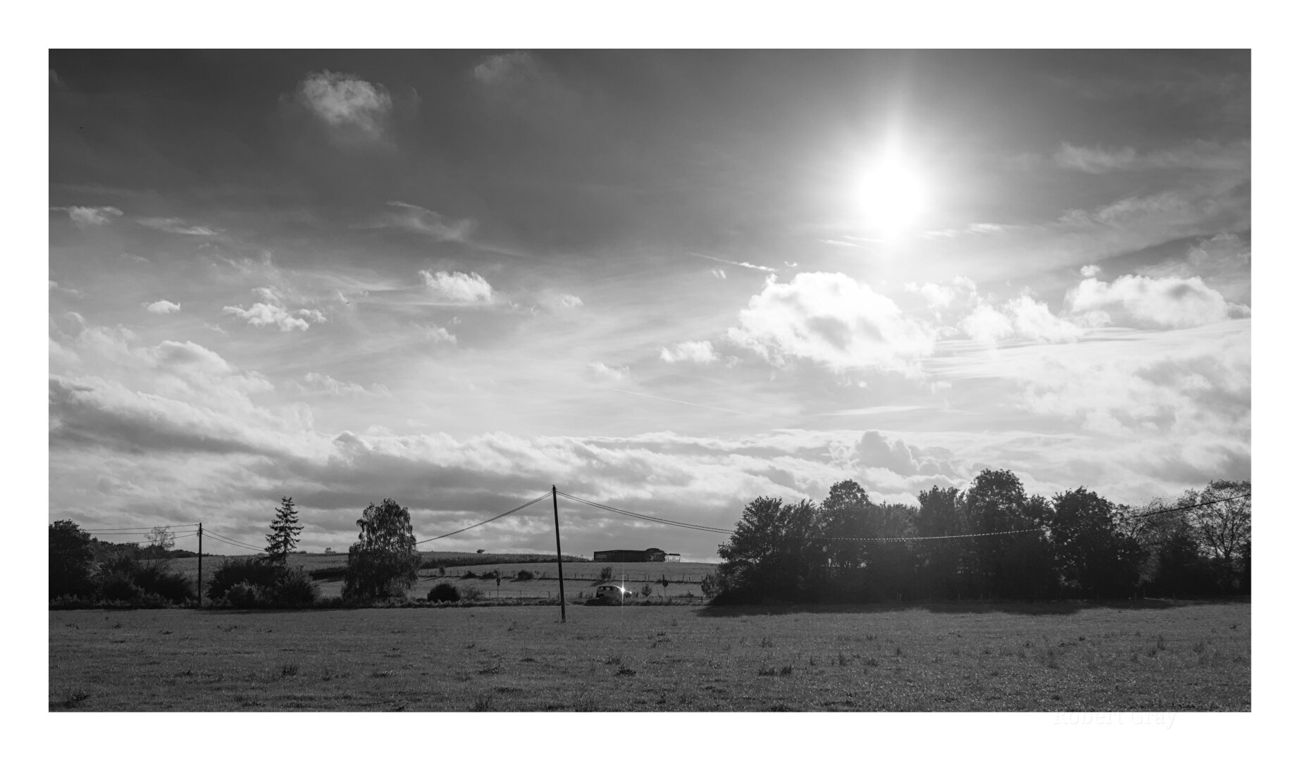 a 16:9 cropped image of a distant road running from left to right, bordered by telegraph poles and wires and trees. In the foreground is an empty meadow. The shot is framed by an imposing and dark sky. The sun dominates the top right of the frame. The image is in high contrast monochrome.