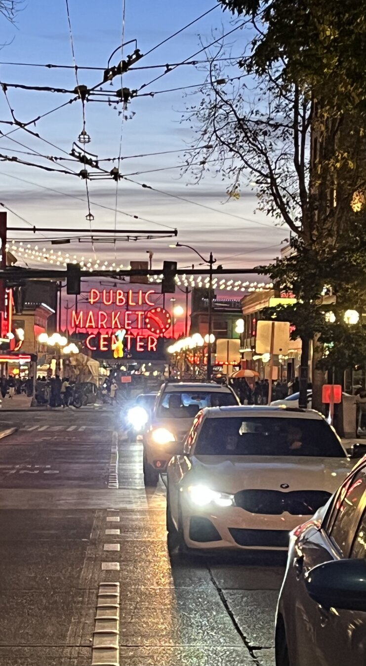 Cars in foreground, headlights on at twilight; Pike Place Market lit up in background.