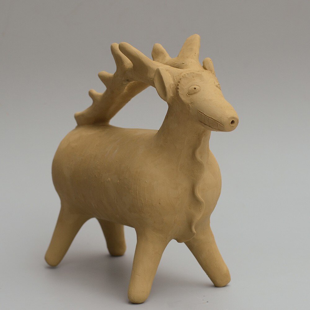 Grey background with an ochre colored vessel in the middle that looks like a stag on four legs. The body is barrel shaped, the face has a tiny hole in the front of the snout and the antlers reach from the head towards the tail, forming a handle by which it can be picked up.