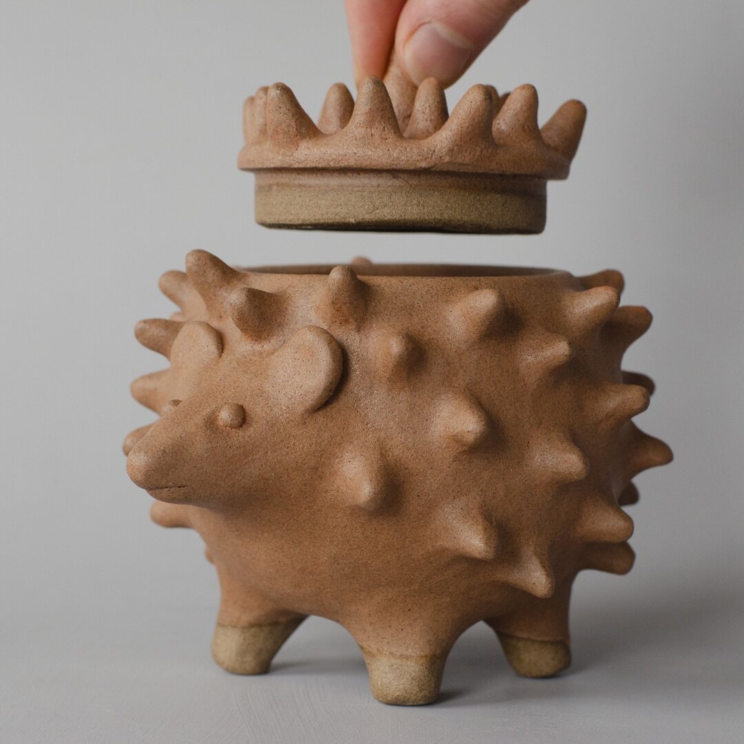 neutral grey background with a sculpture of a brown hedgehog on four legs with a lid that is being lifted by two fingers.