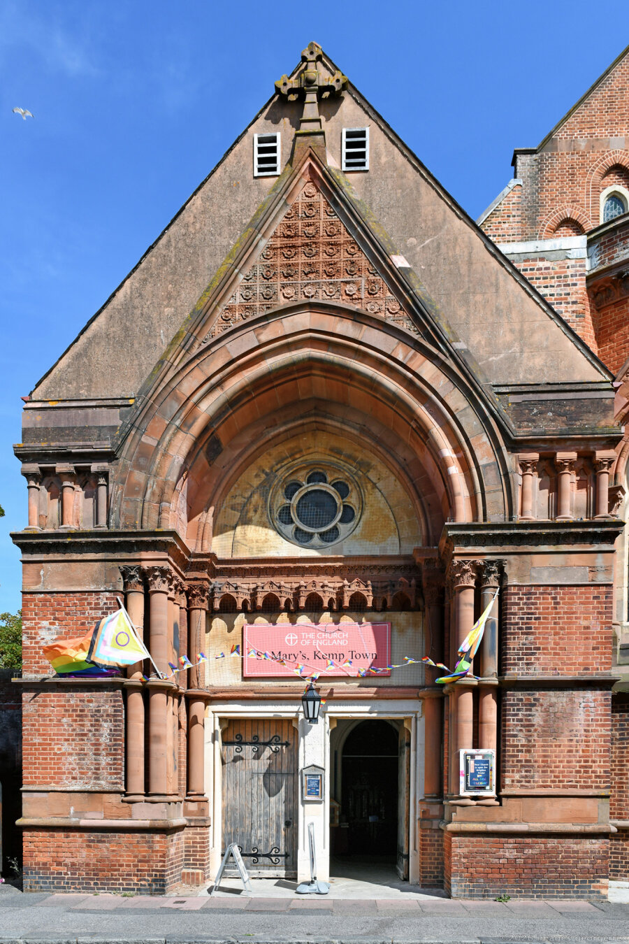 Photo of a Victorian church built in a Gothic Revival style using red brick with pink sandstone dressings. Here showing the entrance porch at the liturgical 'West Front' although actually facing south. Grade 2* listed, City of Brighton & Hove.
