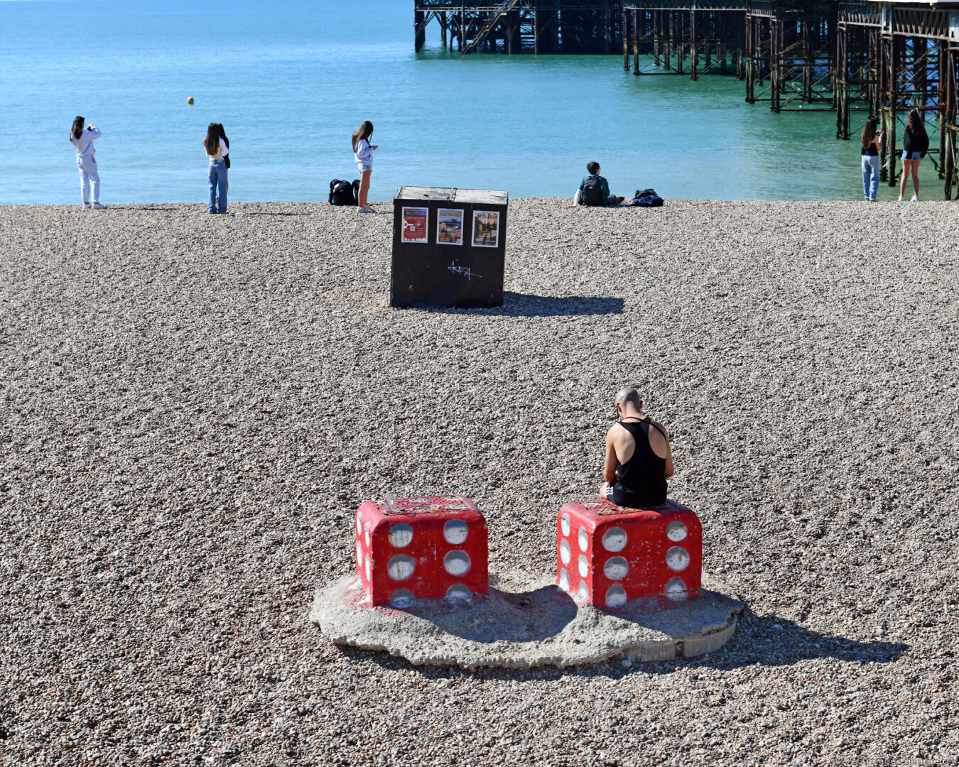 Photo of Brighton beach to the east of the Palace Pier, with visitors sightseeing and taking photos. In foreground someone is sitting on one of a pair of enormous red dice.