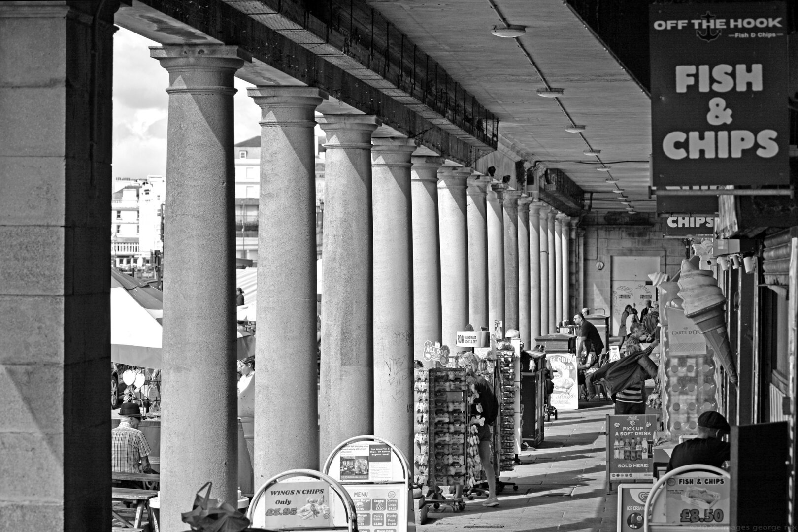 Monochrome photo of The Colonnade on Brighton's Marine Parade. Now mainly occupied by vendors of snacks and souvenirs, plus at the far end the Volks night club.