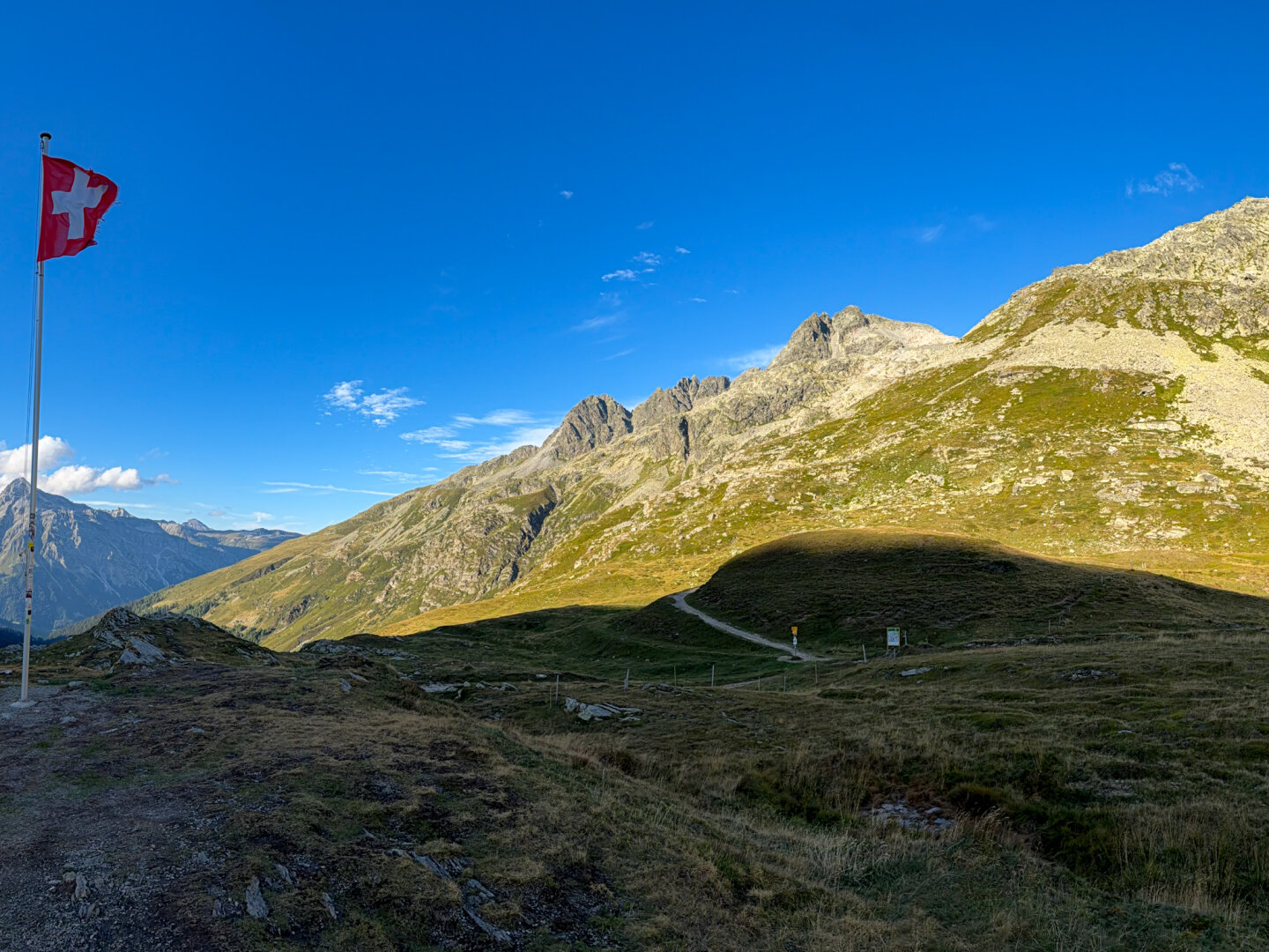 Summit of Splügen Pass looking toward Switzerland. A broad alpine landscape under a bright blue sky with a winding dirt path below rugged mountain peaks. The red and white Swiss flag flies from a tall pole on the left.